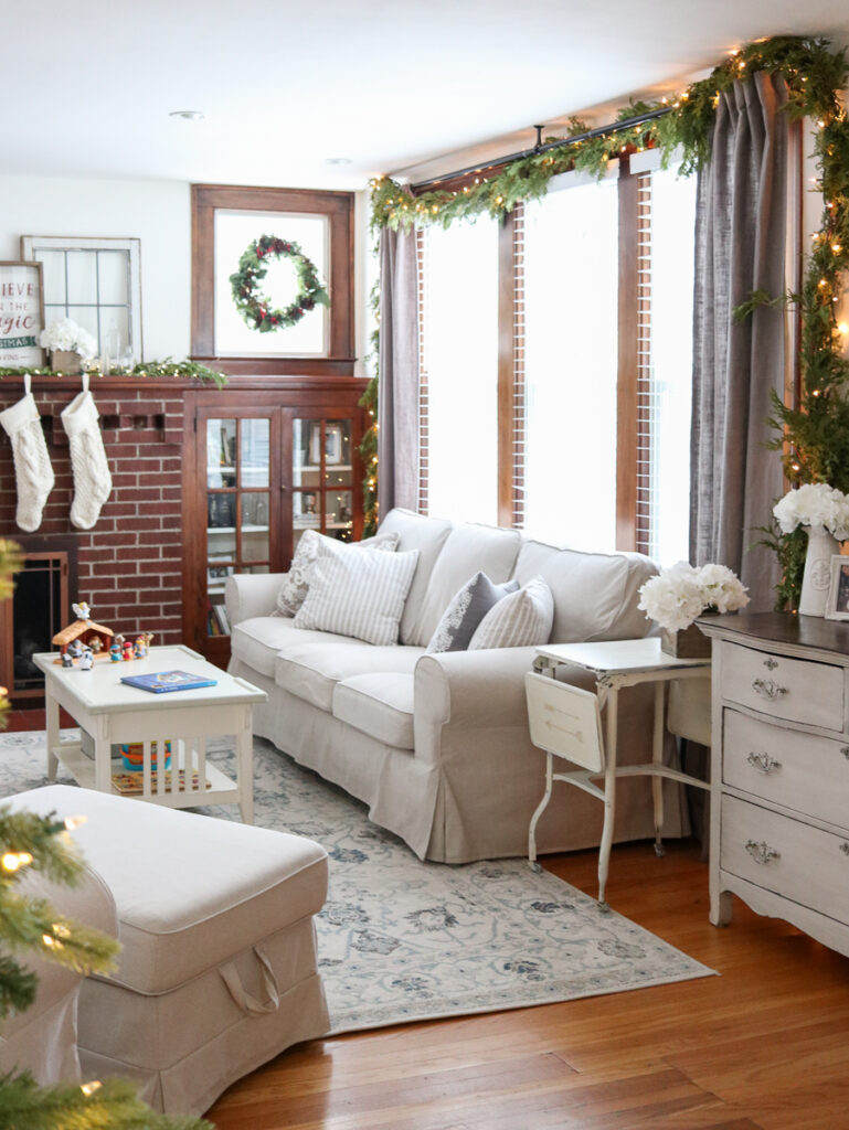 Looking for some neutral Christmas decor inspiration? Stop by to take a peek at our cozy living room decorated for the holidays. Our 1920s living room decor features hardwood floors, built-in cabinets around the mantle with simple fresh greenery and warm white LED lights. Ivory knit stockings adorn the fireplace to complete the look, which tie pin perfectly to the light beige slipcovered sofa, chair and ottoman from the Ikea Ektorp line. 