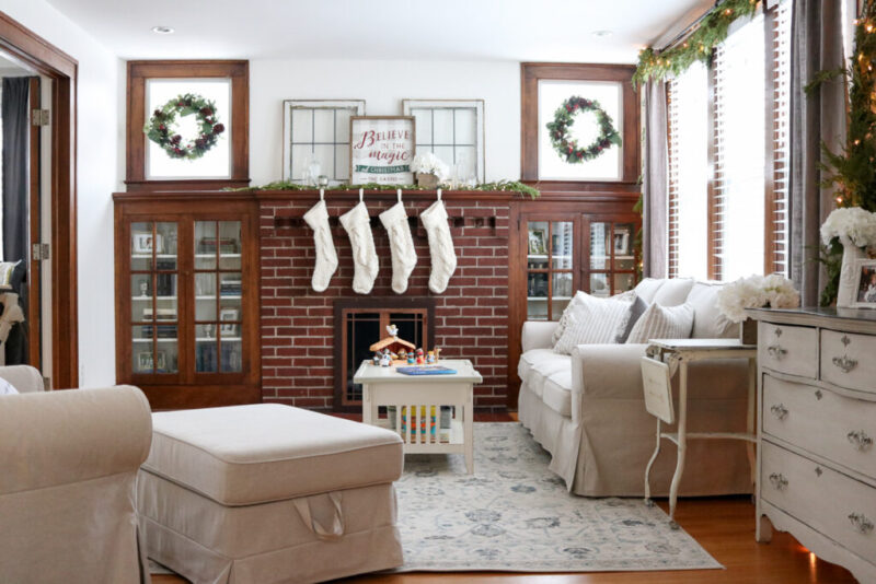 Looking for some neutral Christmas decor inspiration? Stop by to take a peek at our cozy living room decorated for the holidays. Our 1920s living room decor features hardwood floors, built-in cabinets around the mantle with simple fresh greenery and warm white LED lights. Ivory knit stockings adorn the fireplace to complete the look, which tie pin perfectly to the light beige slipcovered sofa, chair and ottoman from the Ikea Ektorp line. 